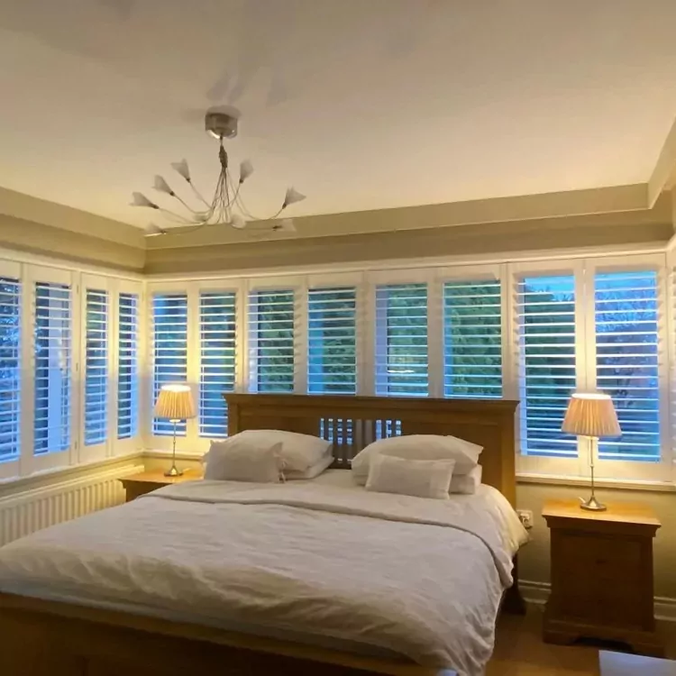 Bedroom with bed, bedside lamps and white plantation shutters around the corner windows