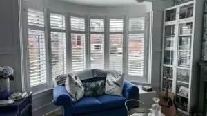 White bespoke shutters installed on curved bay windows in a North Belfast living room with blue sofa and panelled walls.Beautiful Bay Window Shutters in North Belfast Home