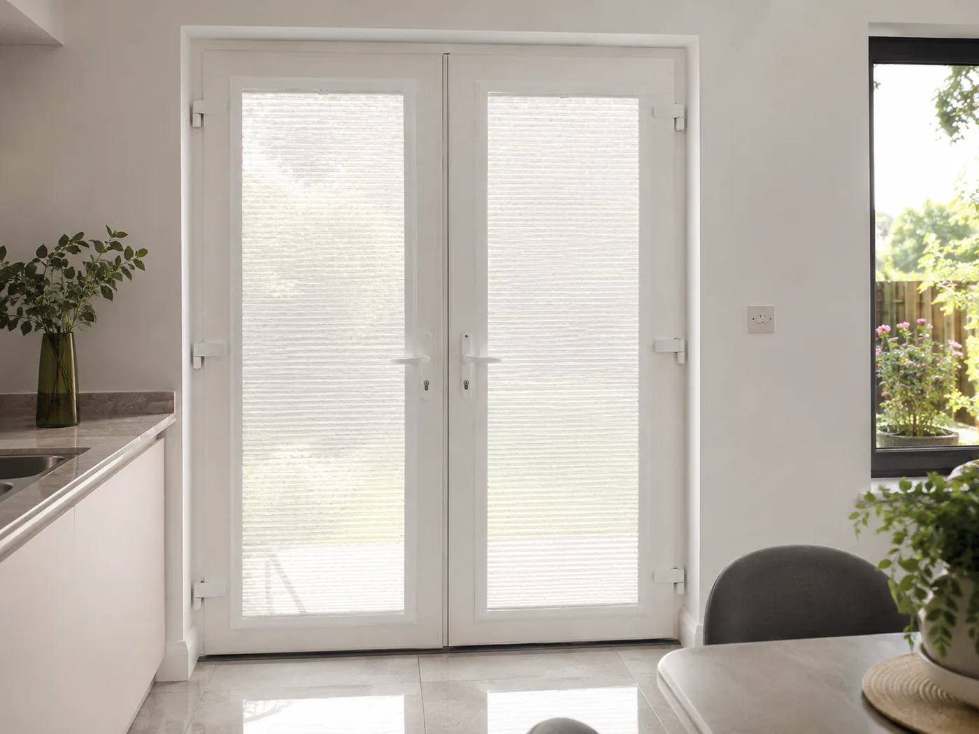 Pale cellular Perfect Fit blinds fitted into white French-door frames in a bright kitchen overlooking a spring garden