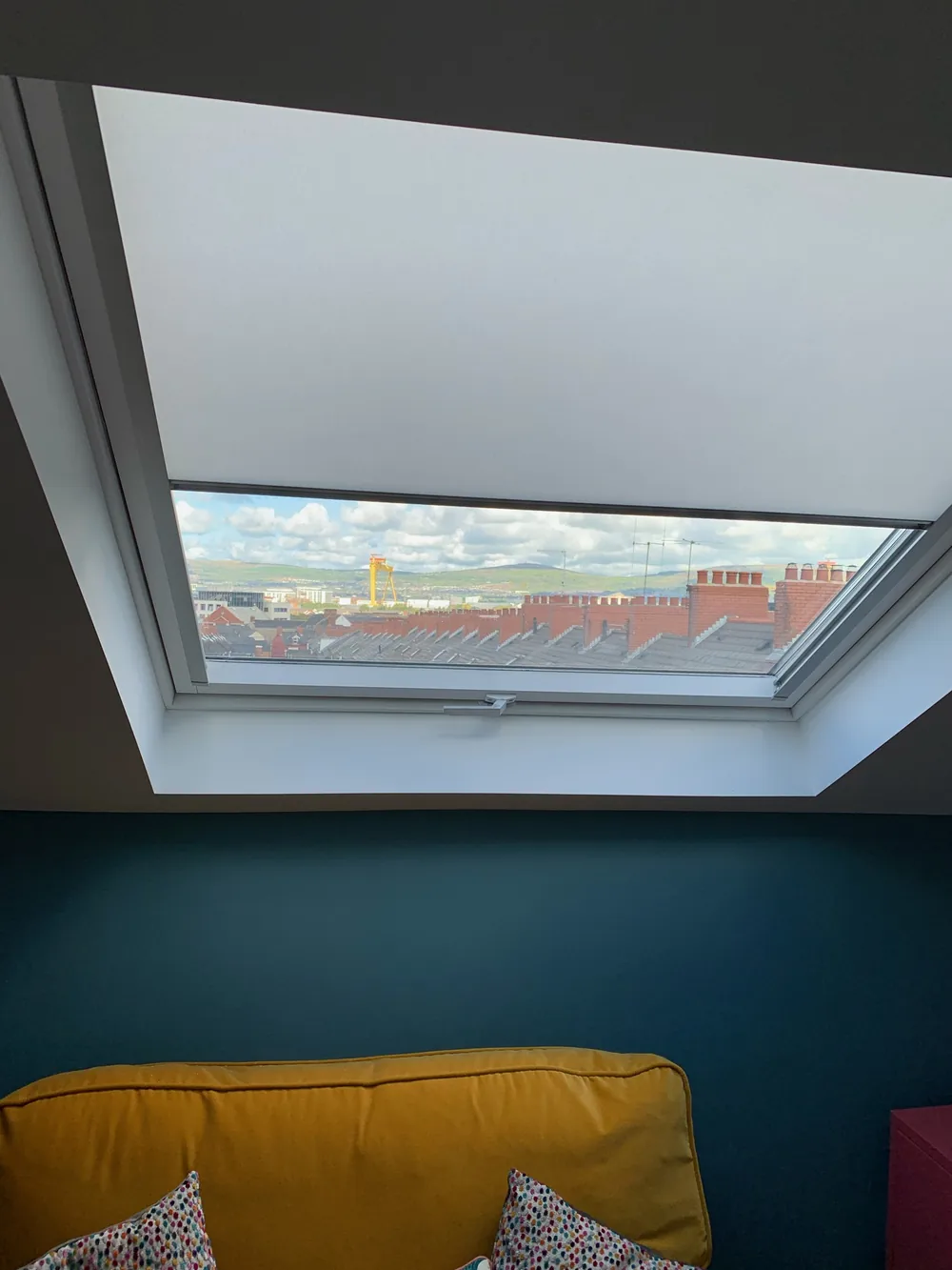 Grey Roller roof window blind partly lowered above a mustard sofa in a colourful attic living room