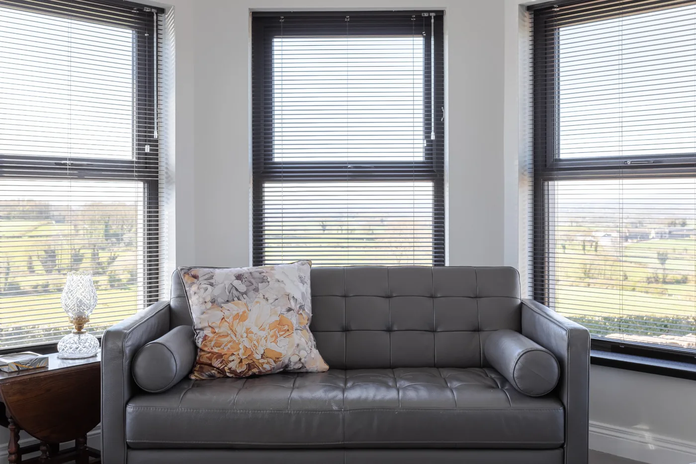 Dark aluminium Venetian blinds fitted to three large living-room windows behind a grey sofa